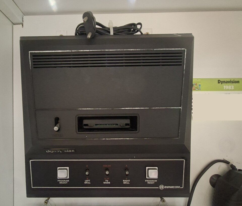 Console Dynavision original de 1983, preto com frisos prateados, entrada de cartucho frontal e painel com chaves Program Select, Left Skill, TV Mode e Right Skill